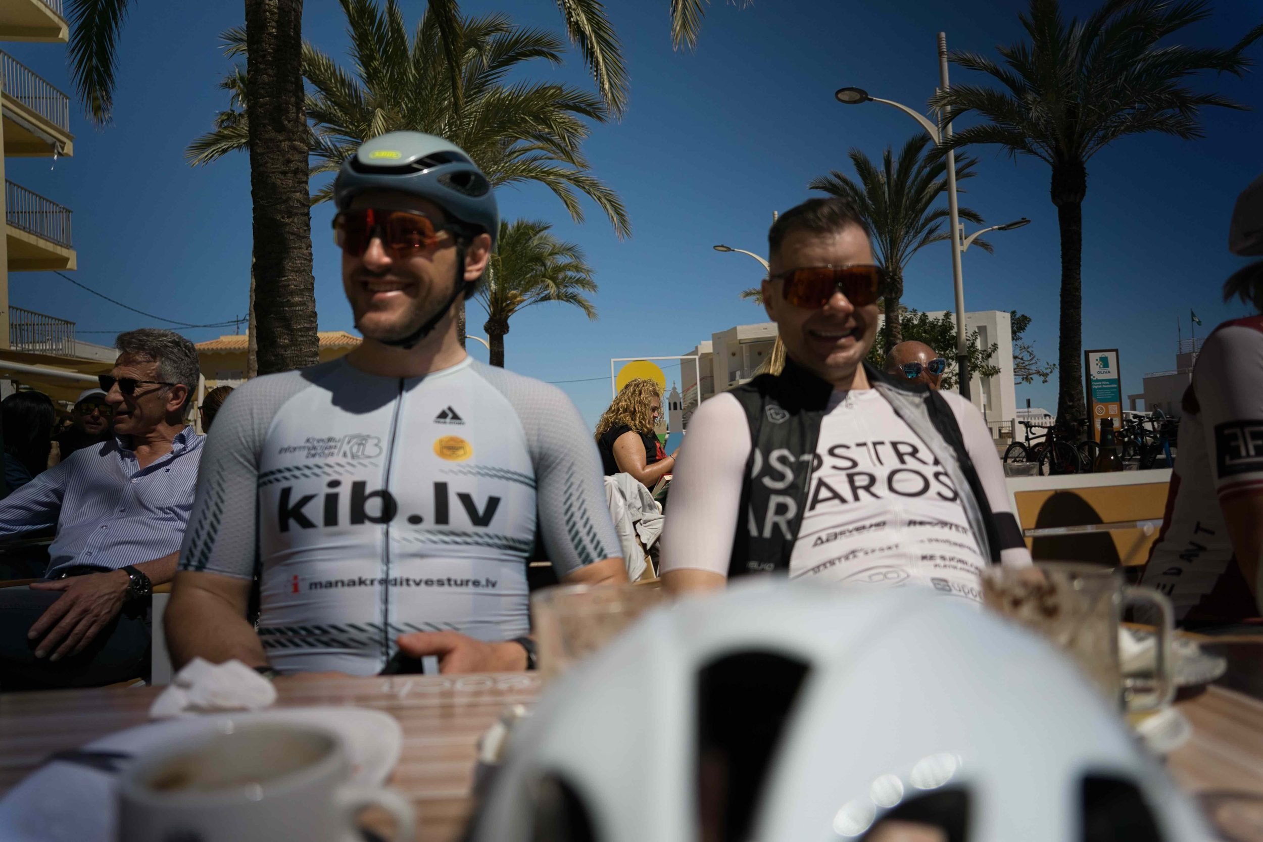 Two Train2Win camp riders smiling at a seaside coffee stop under palm trees