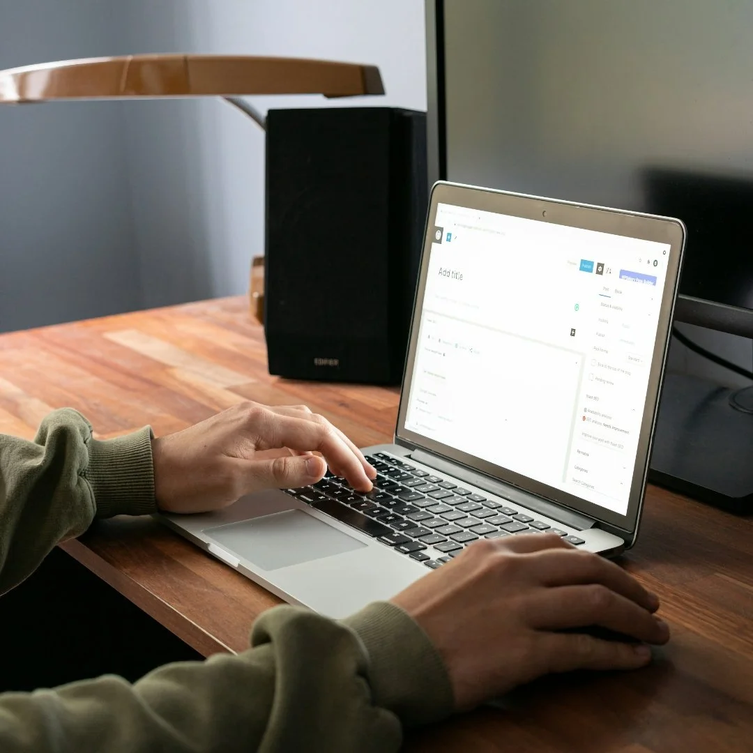 A person working on a silver laptop at a wooden desk with a black speaker and a monitor.