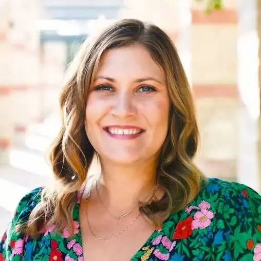 A woman with shoulder-length wavy brown hair, wearing a colorful floral top, smiling outdoors with a blurred background of buildings and trees.
