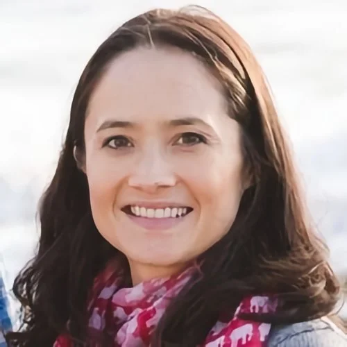 A woman smiling outdoors with snowy mountains in the background.