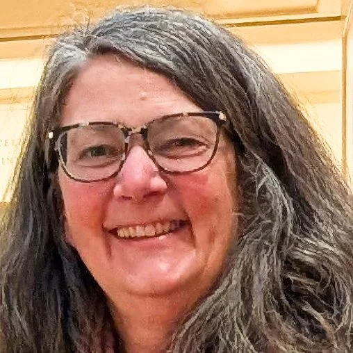 Close-up of a smiling woman with glasses and long, wavy gray hair in a wooden-paneled room.