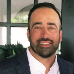 A man with a beard and short dark hair, wearing a navy blazer and white shirt, smiling indoors near a window with green plants in the background.