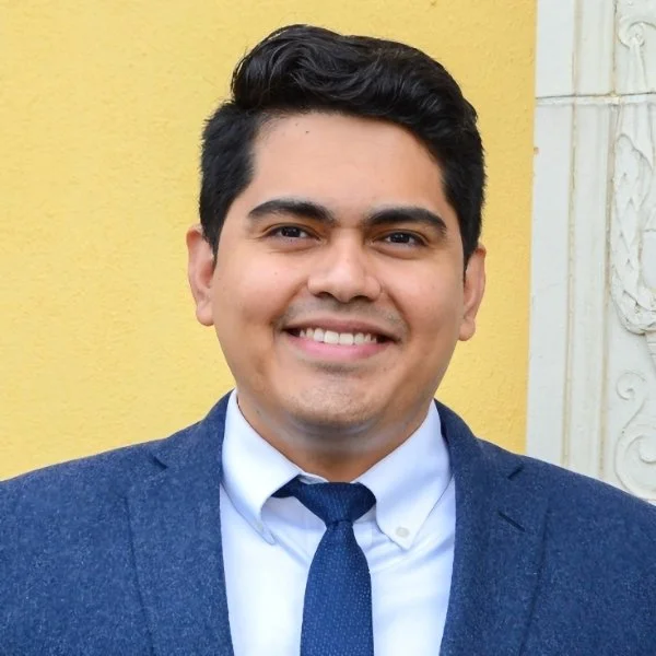 Portrait of a smiling man in a navy suit, white shirt, and navy tie, standing outdoors in front of a light yellow wall.