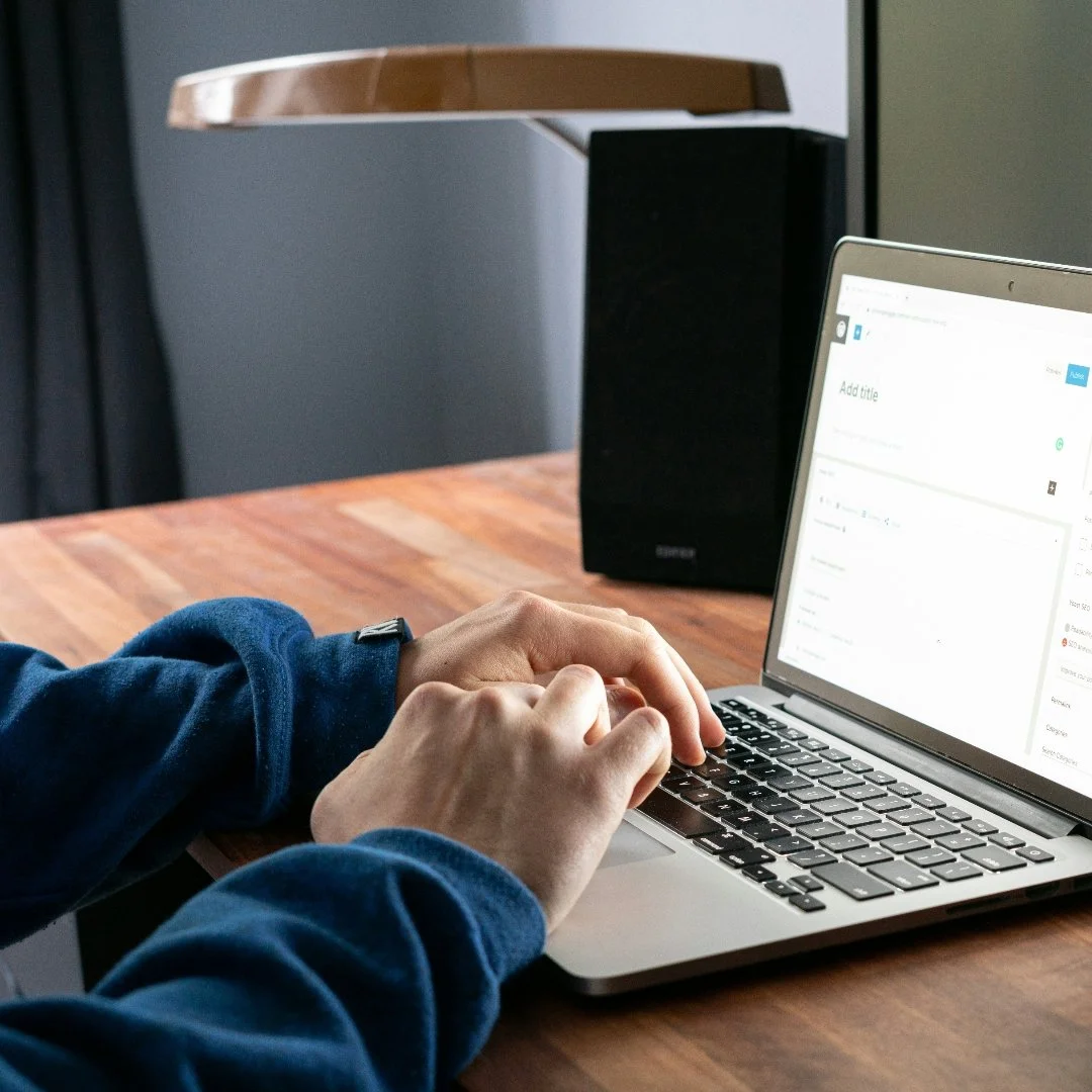 Person typing on a silver laptop with a black speaker and wooden desk in the background.