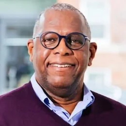 Close-up of smiling man with glasses, short gray hair, wearing a navy sweater over a light blue collared shirt.