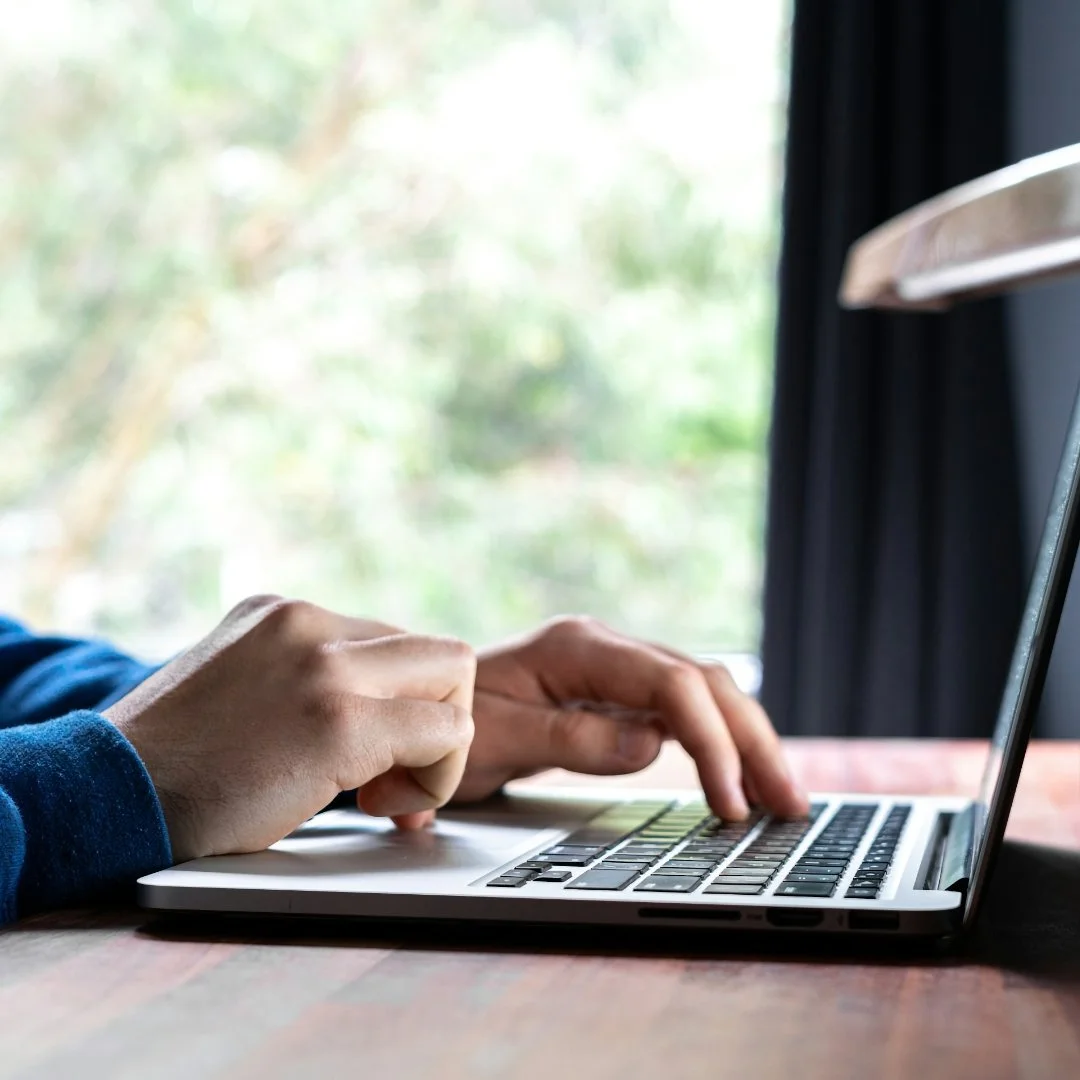 Close-up of person typing on a laptop keyboard at a wooden desk indoors with a window showing blurry trees outside.