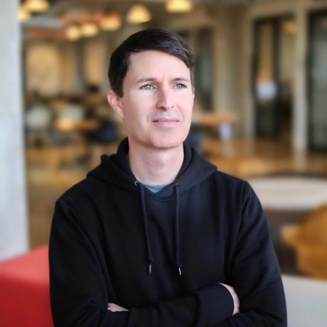 A man with dark hair and light skin wearing a black hoodie, looking thoughtfully to the side in a modern indoor setting.