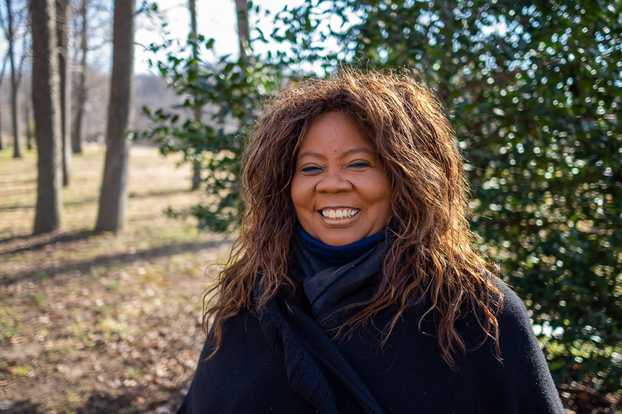 A woman with curly hair smiling outdoors in a wooded area with sunlight shining through trees.