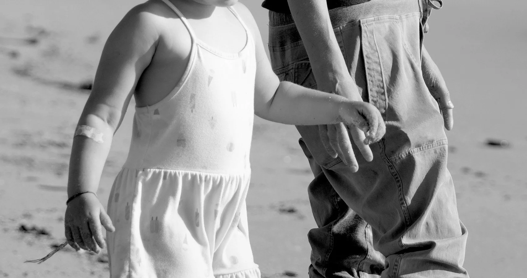 A black and white photo of a child and an adult holding hands on a sandy beach.