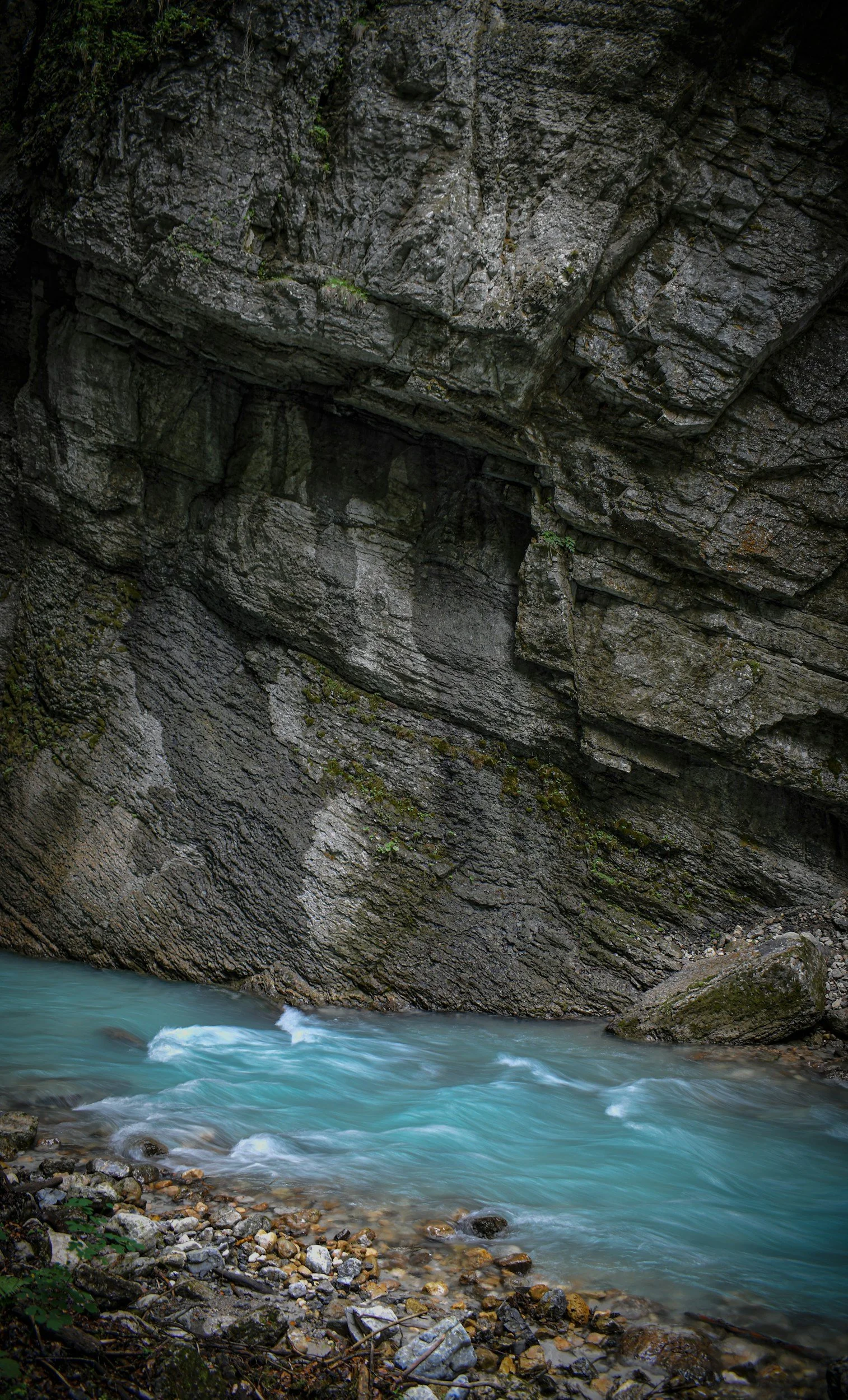 Stream flowing at the base of a large rocky cliff with moss and small plants growing on the rocks.