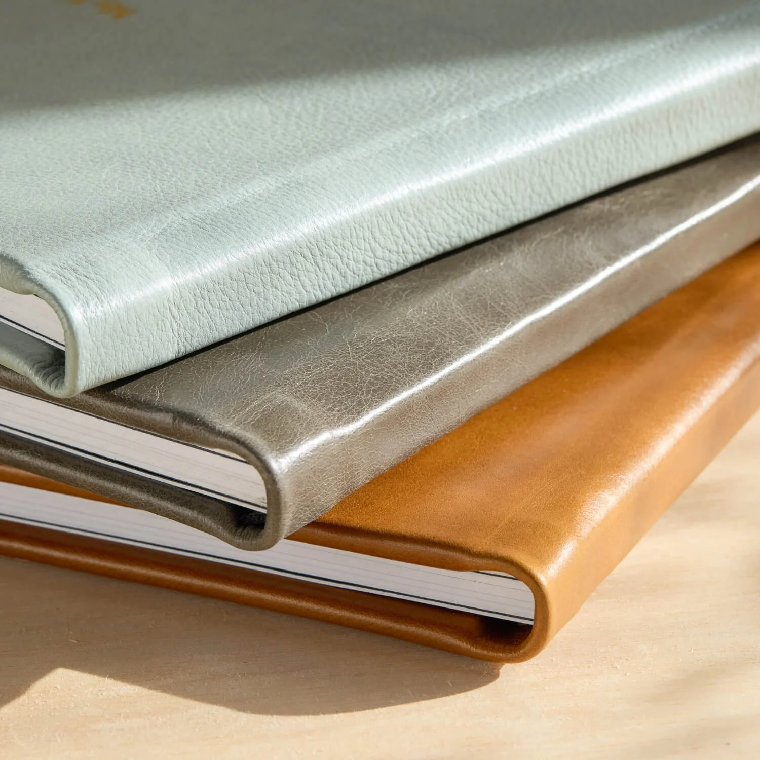 Close-up of three stacked leather-covered notebooks in light green, metallic gray, and brown, on a wooden surface.