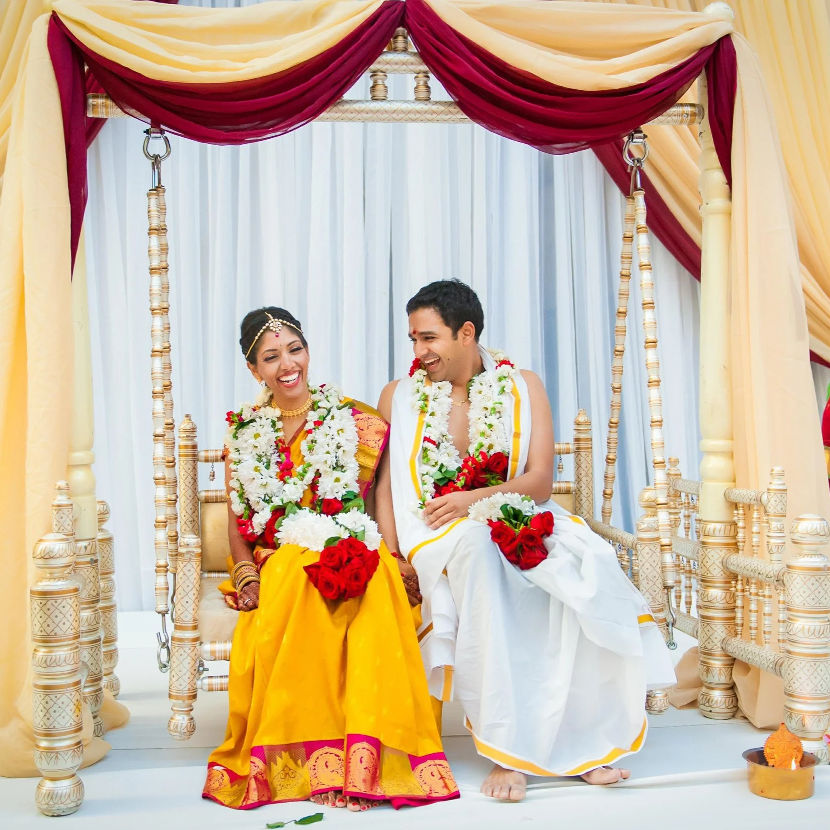 A joyful couple dressed in traditional Indian attire during their wedding ceremony, sitting together on a decorated seat with floral garlands and ornate drapes in the background.