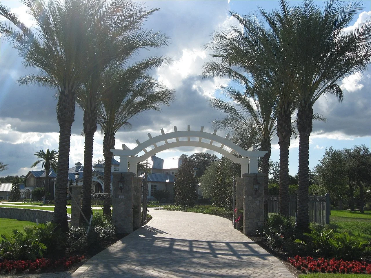 Entrance to a gated community with tall palm trees, a stone and white wood archway, greenery, and houses in the background under a partly cloudy sky.
