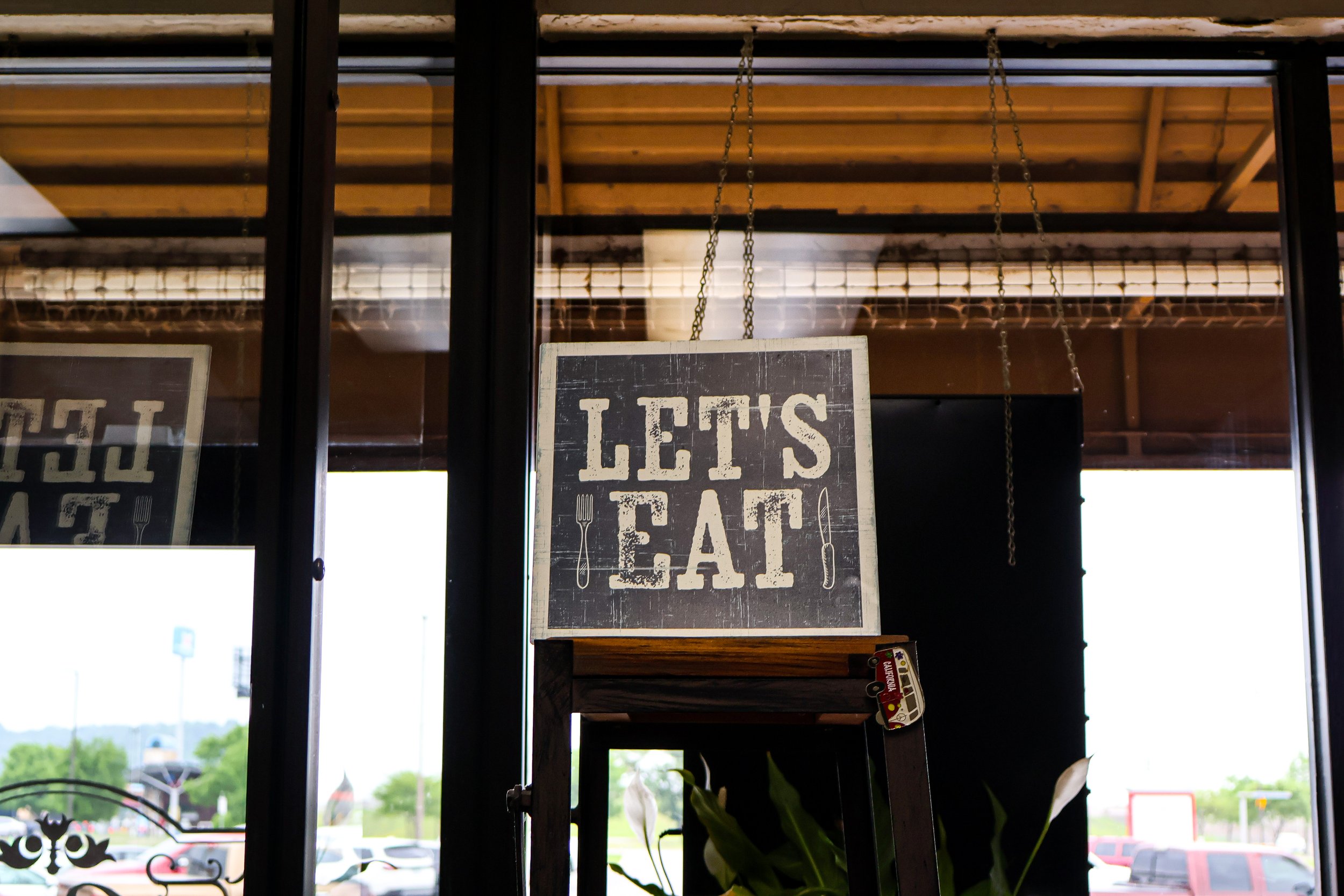 A sign hanging inside a restaurant or cafe that says 'Let's Eat' with a fork on the left and a knife on the right, visible through glass windows during daytime.