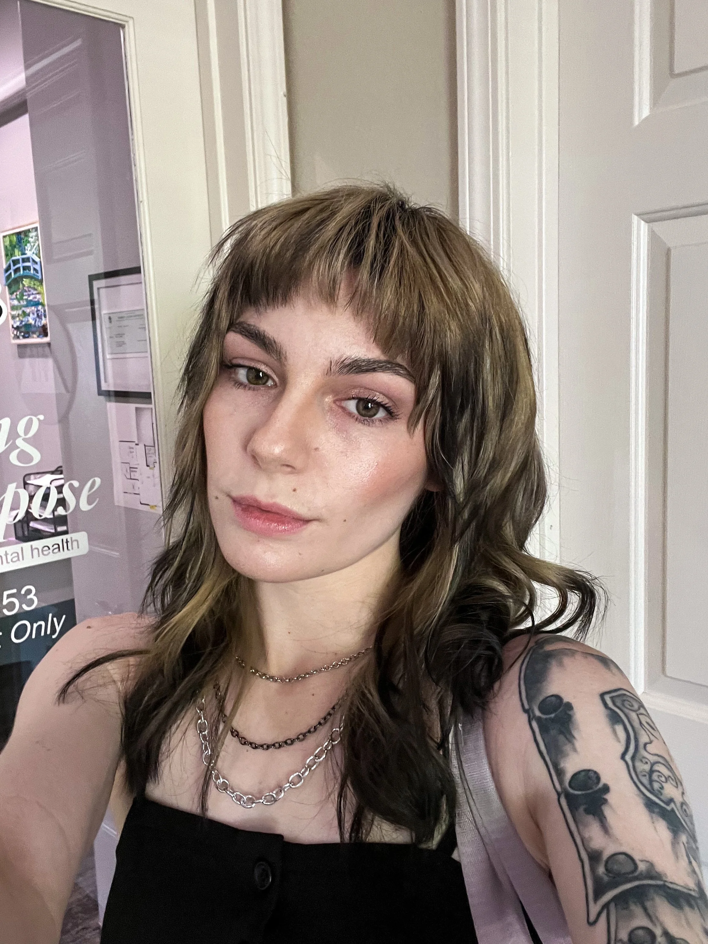 A femme-presenting non-binary person with blonde hair that gradients to black ends. They are wearing a black tank top and taking the photo in a selfie-style. Their expression is a slight smile.