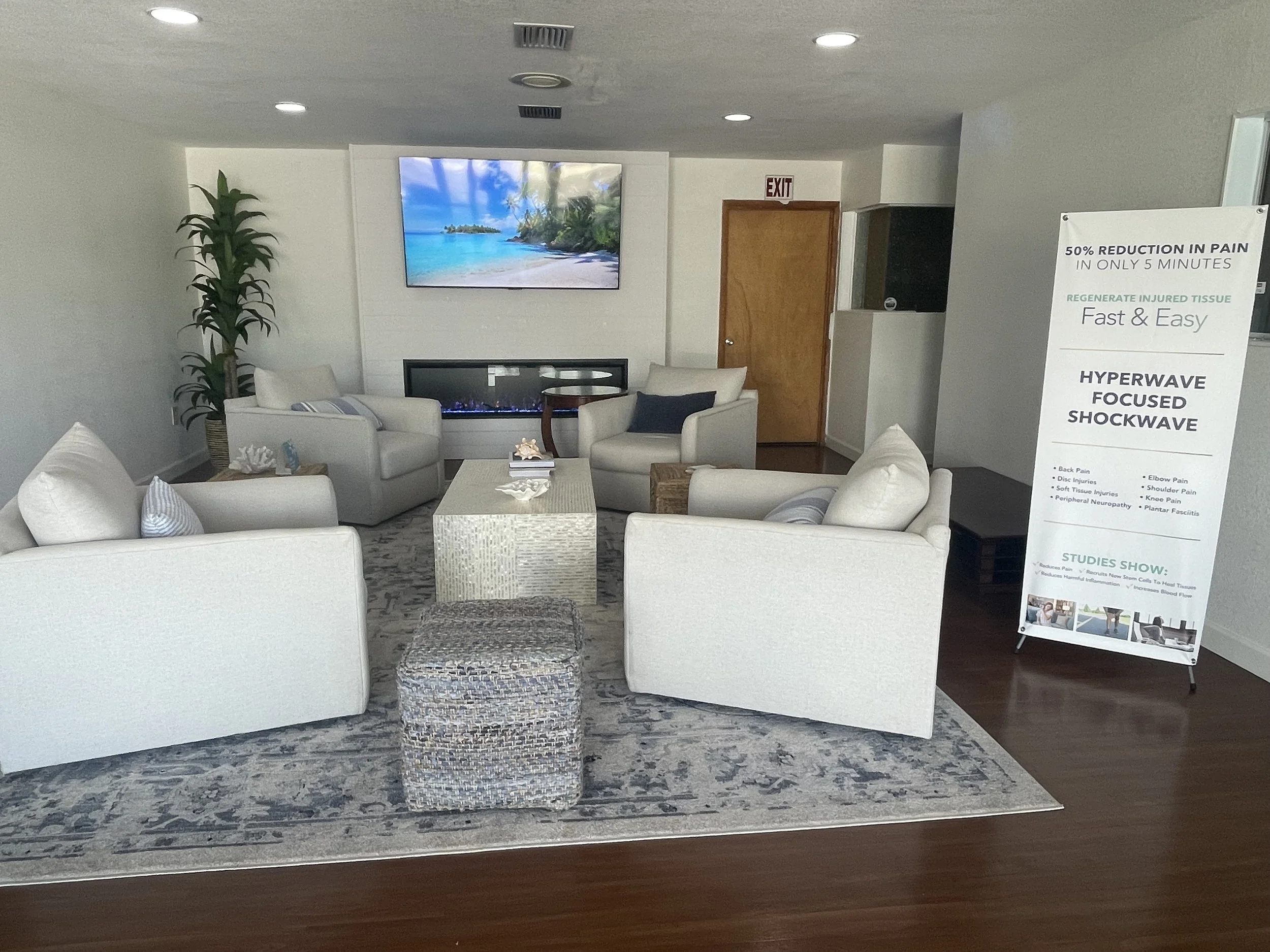 A waiting room with white sofas, a potted plant, a wall-mounted TV displaying a beach scene, and a sign about pain relief on a stand. The room has recessed ceiling lights and hardwood flooring.