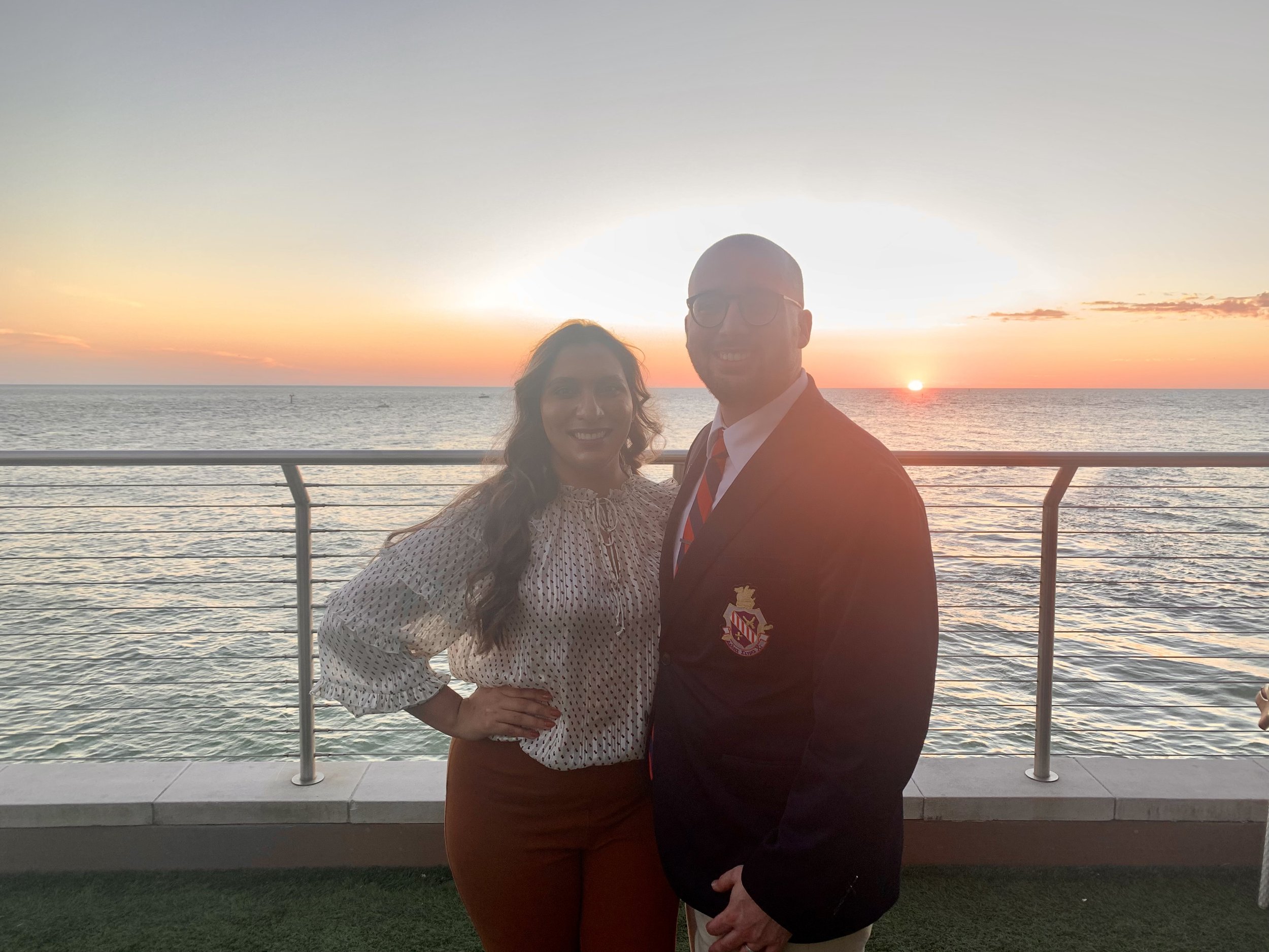 A man and woman standing together on a balcony by the water during sunset, smiling at the camera. The woman has long, wavy hair and is wearing a polka dot blouse and rust-colored pants. The man is wearing glasses, a suit with a crest on the blazer, a white shirt, and a striped tie.