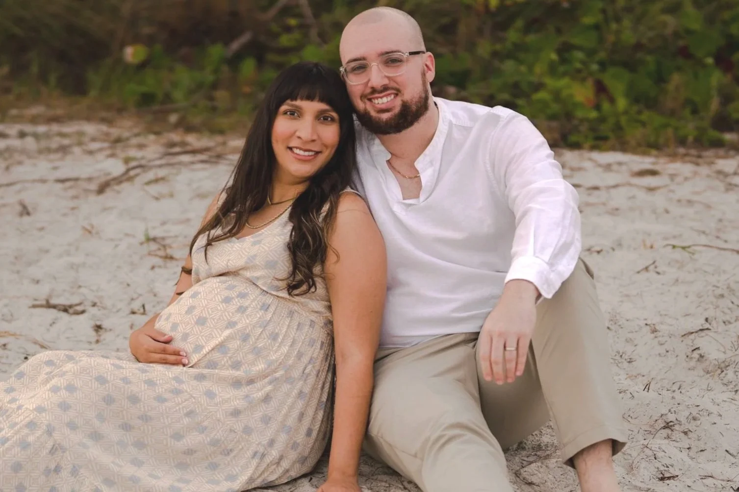 A pregnant woman and a man sitting on the sandy beach, smiling at the camera with green foliage in the background.