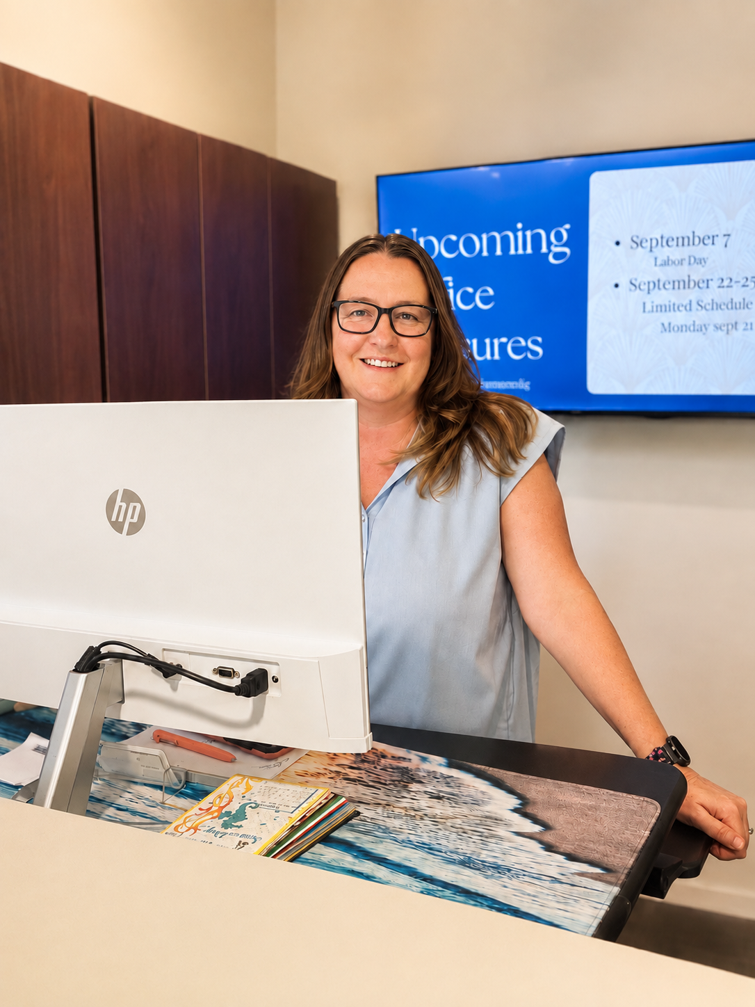 Smiling woman with glasses sitting at a reception desk with a computer, a colorful brochure, and a digital screen displaying upcoming office closures.