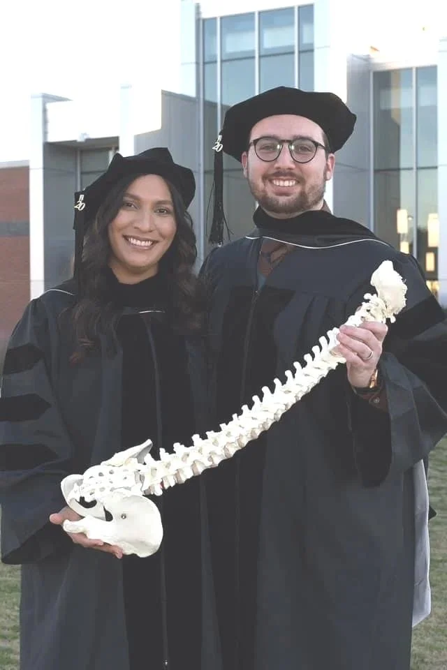Two graduates wearing caps and gowns holding a human spine model, standing outside a modern building, smiling.
