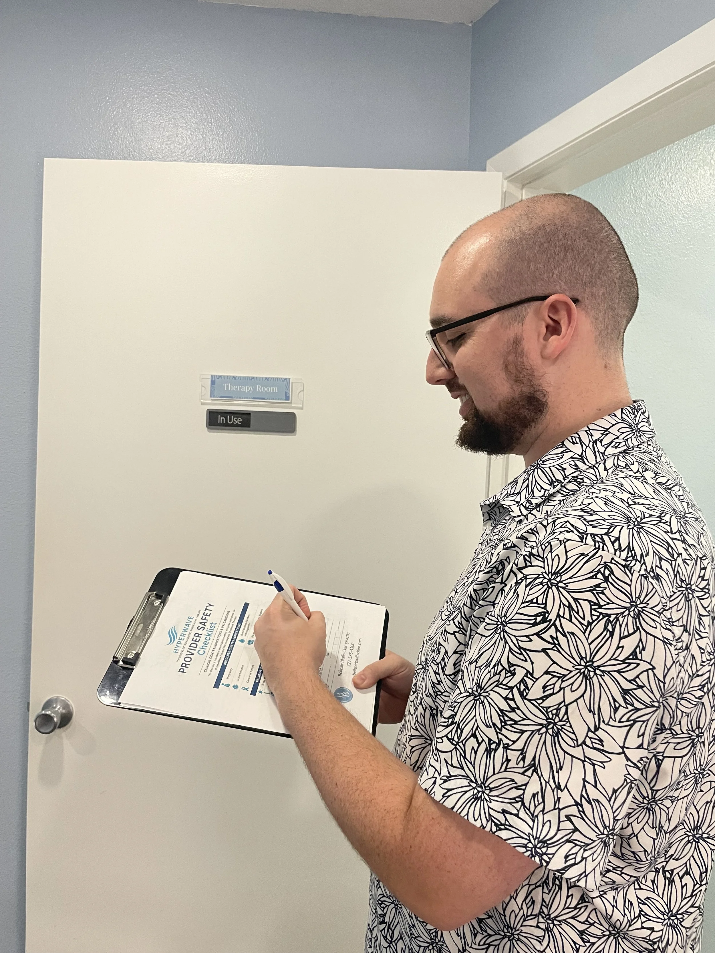 A man with glasses, a beard, and a floral shirt, smiling while filling out a clipboard with a safety checklist in front of a white door labeled 'Therapy Room' and 'In Use' sign.