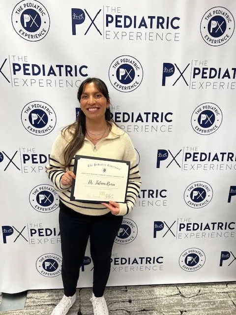 A smiling woman holding a certificate in front of a step and repeat backdrop with logos for The Pediatric Experience and PX.