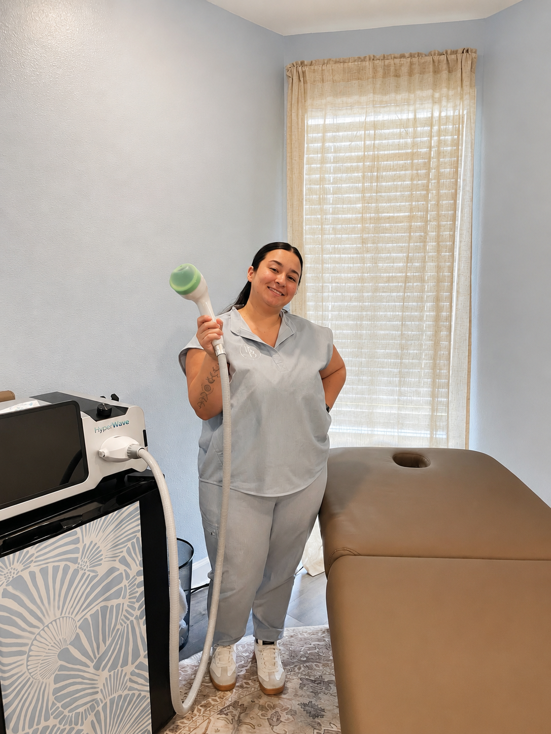 A smiling woman dressed in grey scrubs holding a handheld device in a treatment room with a massage table and a machine labeled 'HyperWave'.