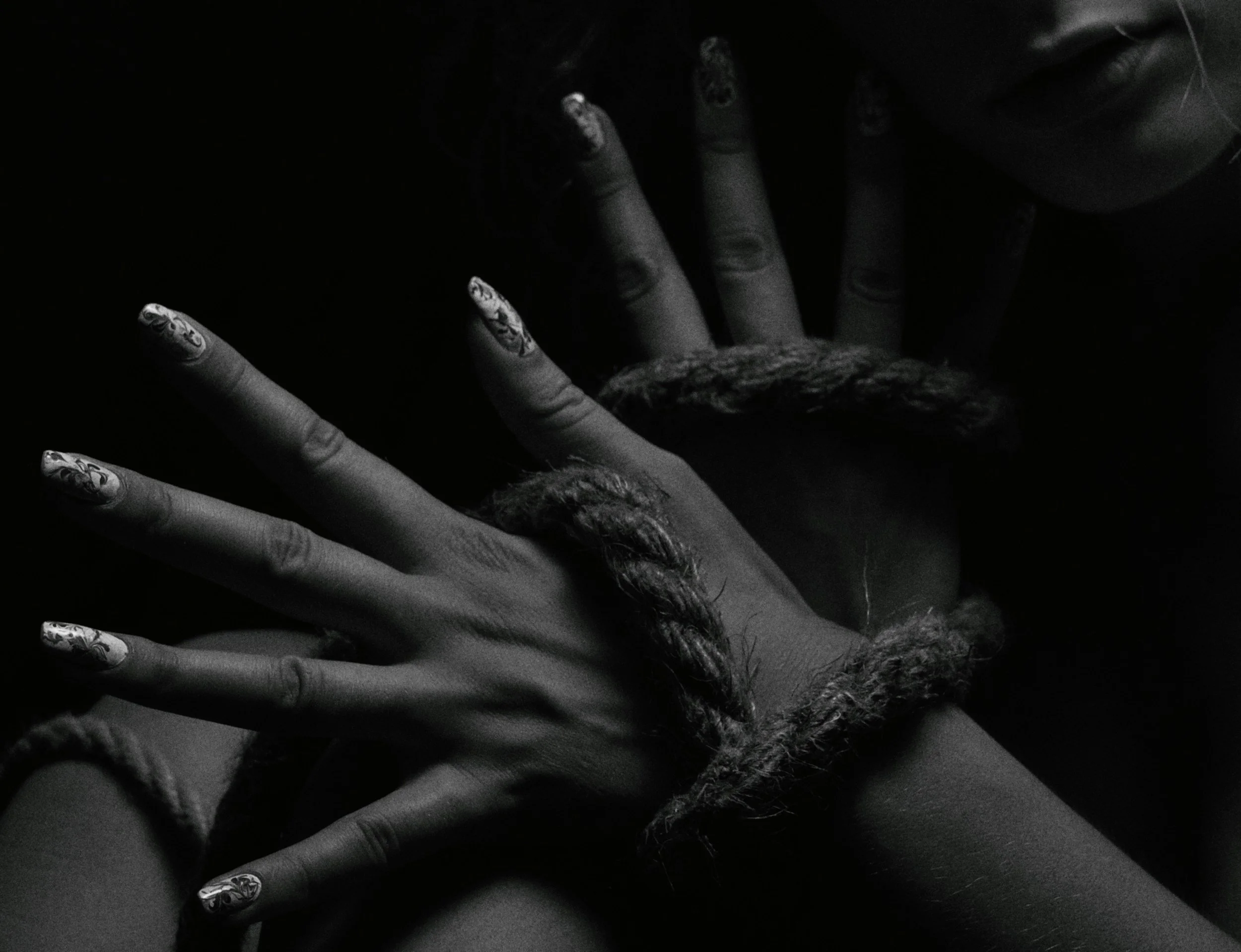 Close-up black and white photograph of two hands intertwined, with one hand having floral painted fingernails and the other with a knitted bracelet, partially covering a person's face.