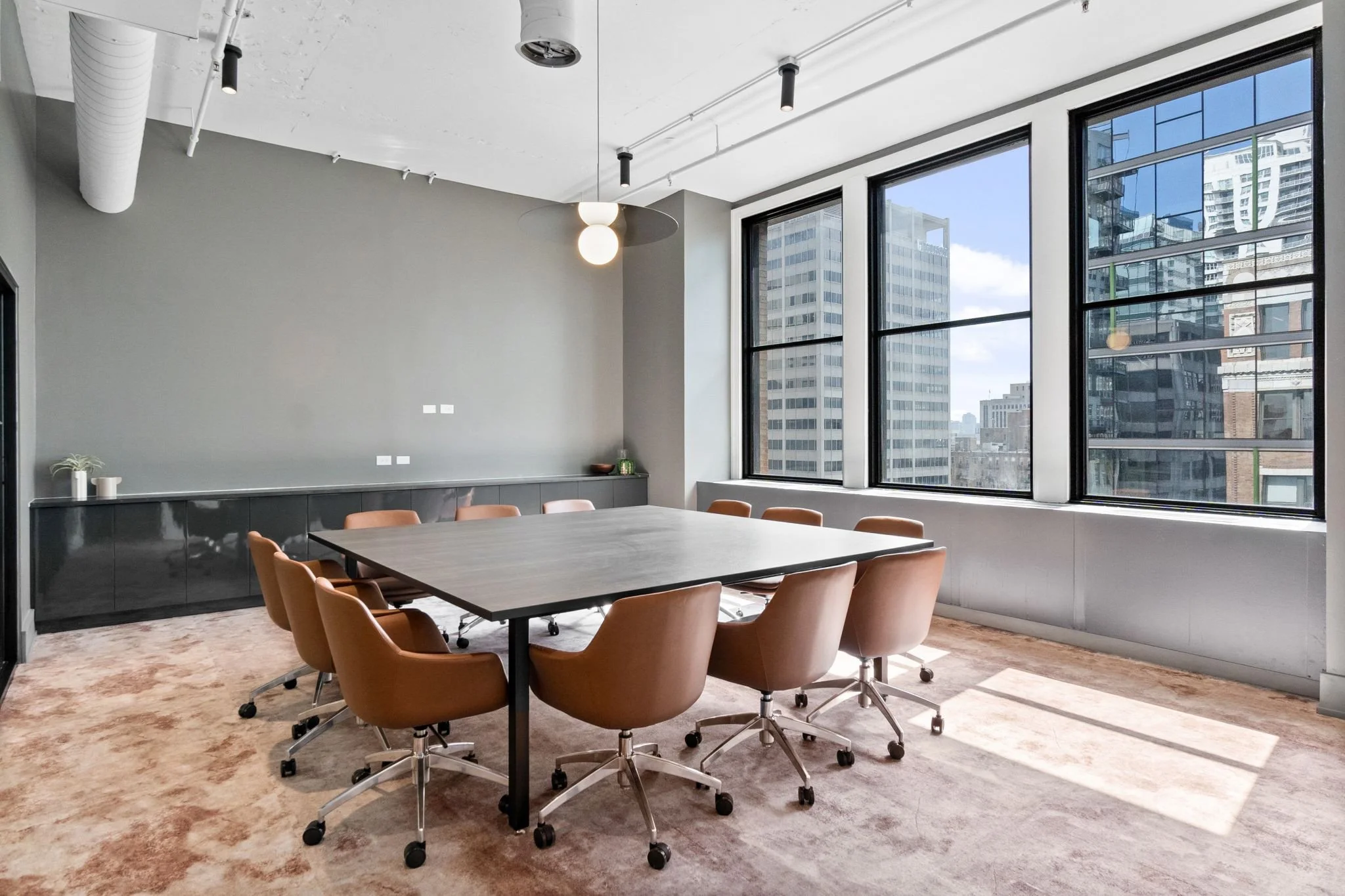 A modern office conference room with a large table surrounded by tan swivel chairs on casters. The room has large windows with a cityscape view of tall buildings, and a gray accent wall with minimal decor, including vases and a small bowl. The ceilin