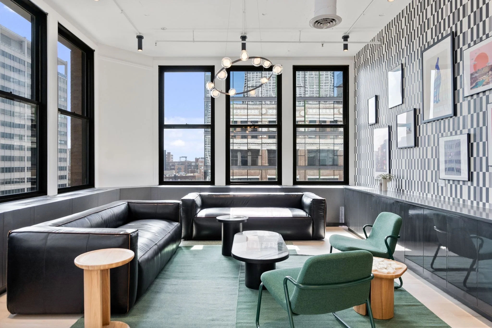Modern office lounge with black leather sofa, green armchairs, round coffee tables, large windows, geometric wallpaper, and artwork on the wall.
