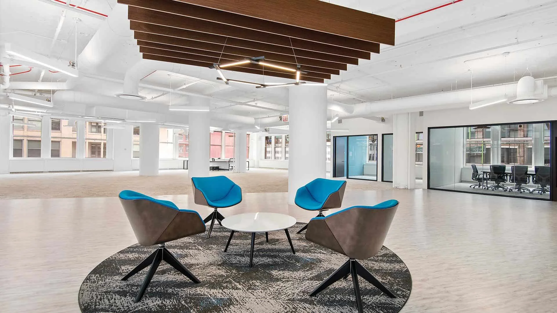 Modern office lounge area with five blue and gray chairs arranged around a small white round table on a patterned rug in the foreground. The background features an open office space with large windows, white columns, and a conference room with glass 