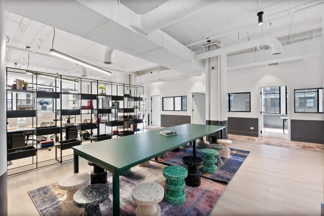 Modern, open-concept office space with large windows, a green table with assorted colorful stools, and a black metal bookcase with books and decorative items.