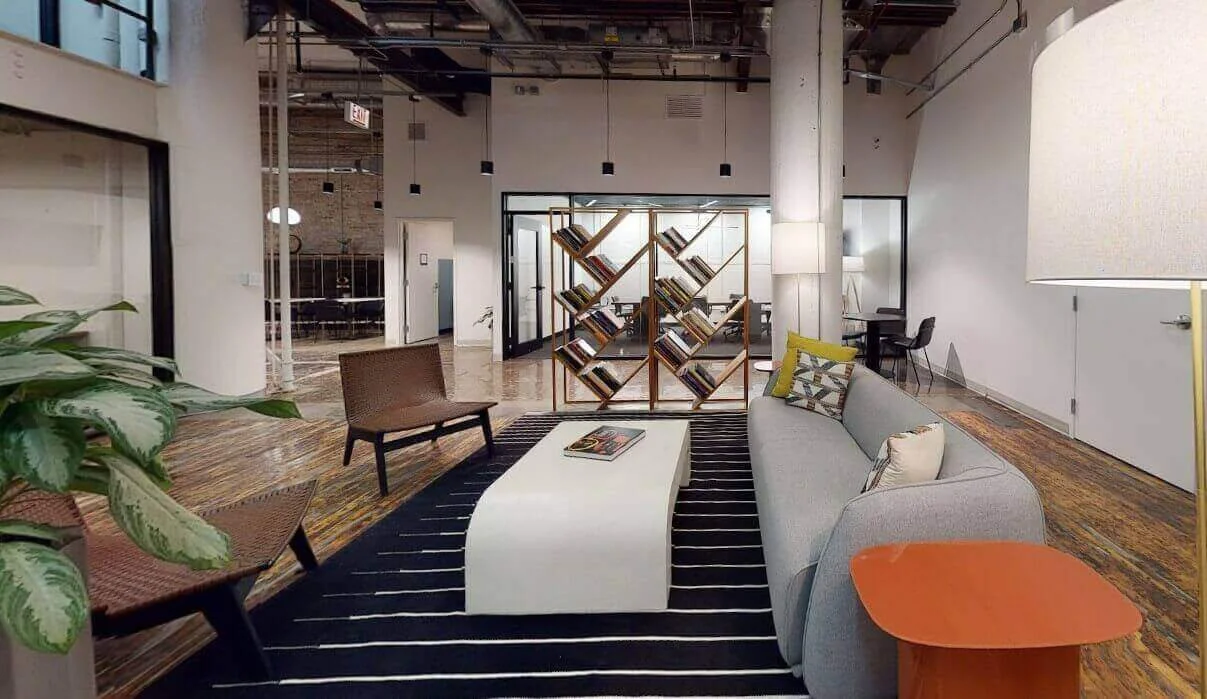 Modern office lounge area with a gray sofa, wooden coffee table, two brown chairs, a tall lamp, a orange side table, a black and white striped rug, a bookshelf, and a workspace with a desk and chair in the background.