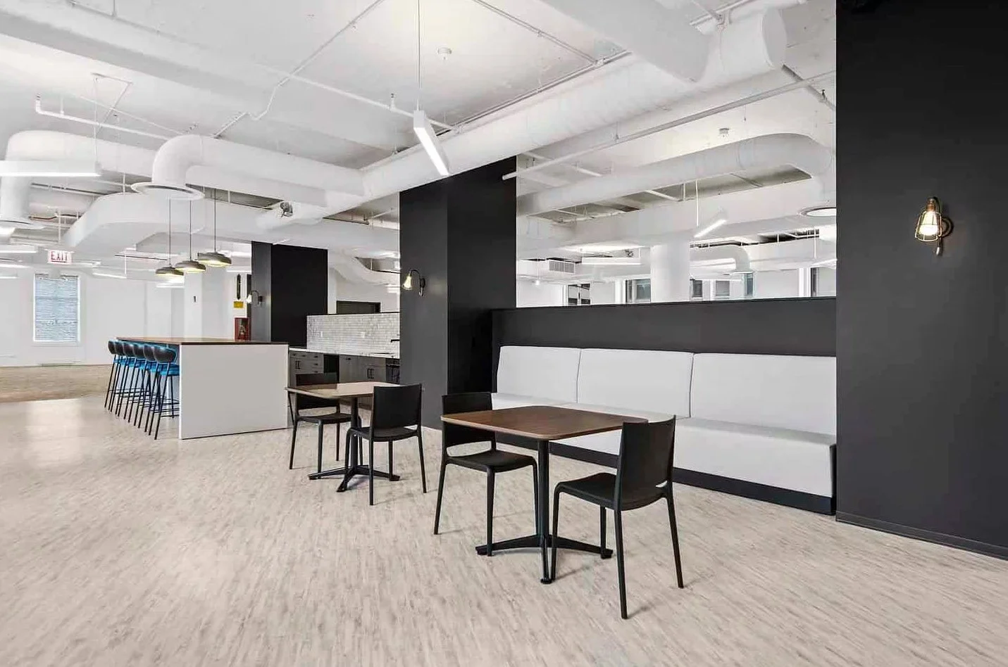 Modern cafeteria with black and white decor, wooden tables, black chairs, lounge seating, exposed ceiling pipes, and wall sconces.