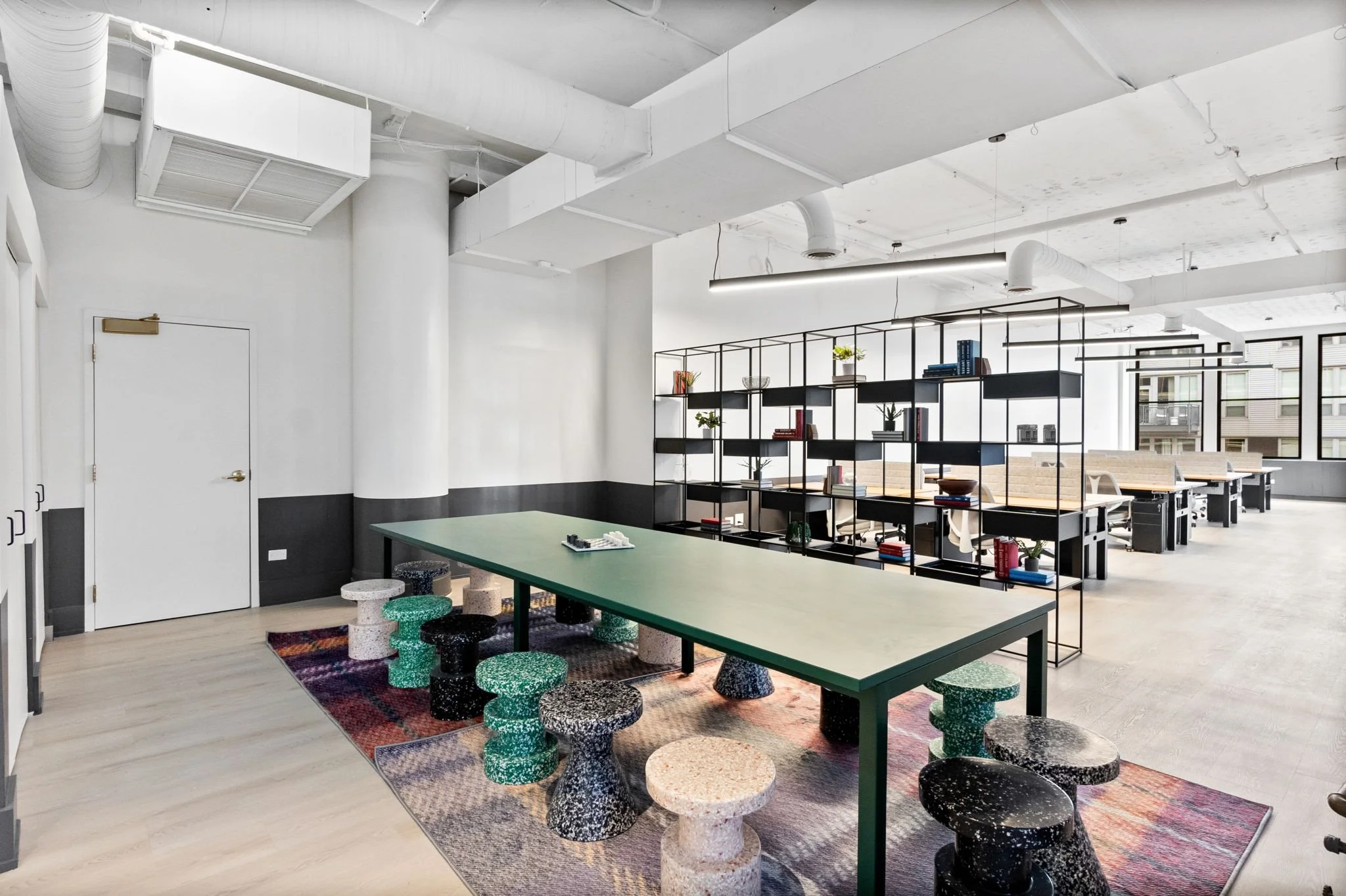 Modern open office space with a green rectangular table surrounded by colorful speckled stools, a black shelving unit with books and plants, and large windows allowing natural light.