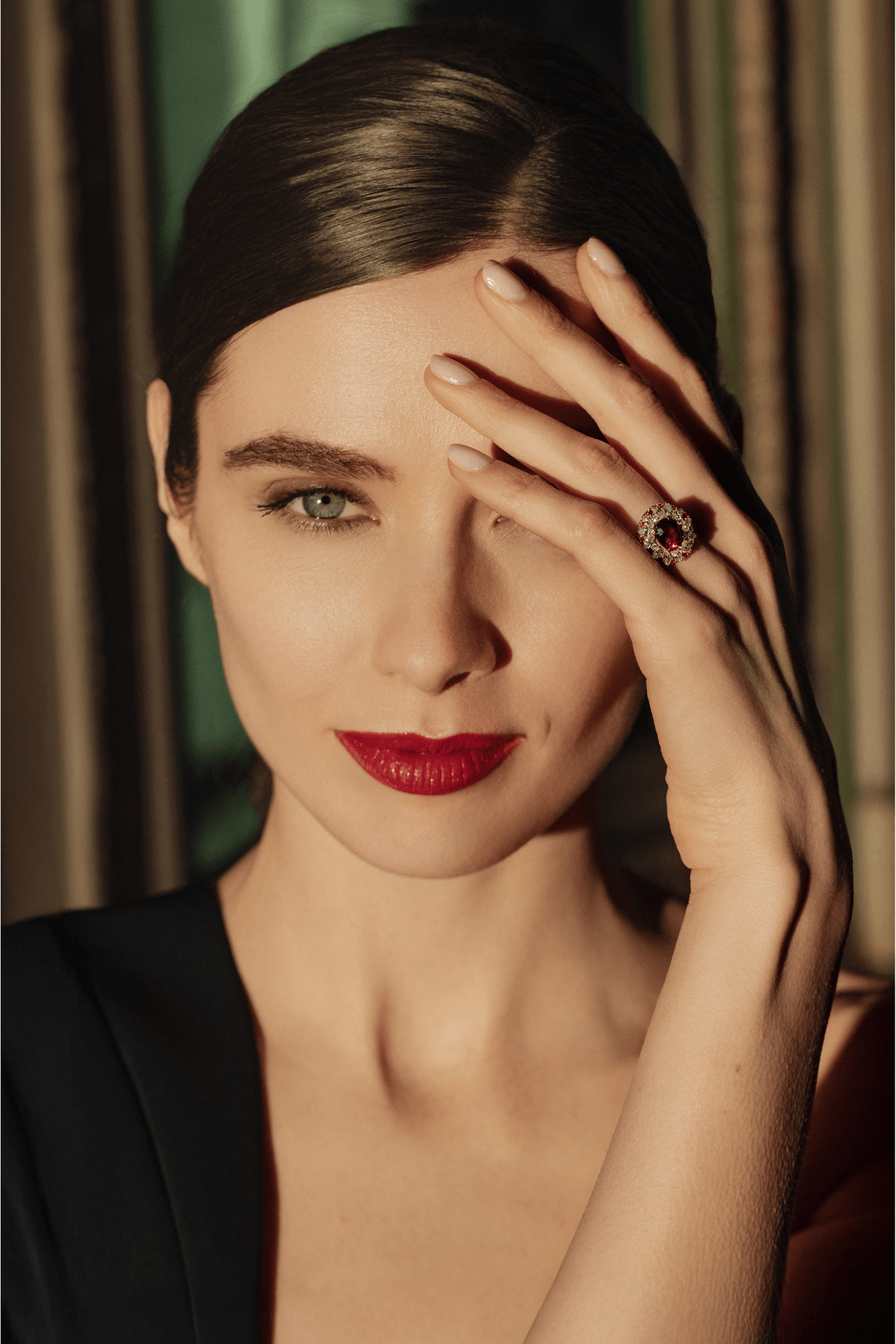 Une femme aux cheveux bruns coiffés élégamment, portant un maquillage sophistiqué avec du rouge à lèvres rouge. Elle porte une bague ornée d'une pierre rouge entourée de diamants, et une robe noire. Elle pose avec une main couvrant partiellement son œil gauche, dans un arrière-plan de rayures vertes et beiges.