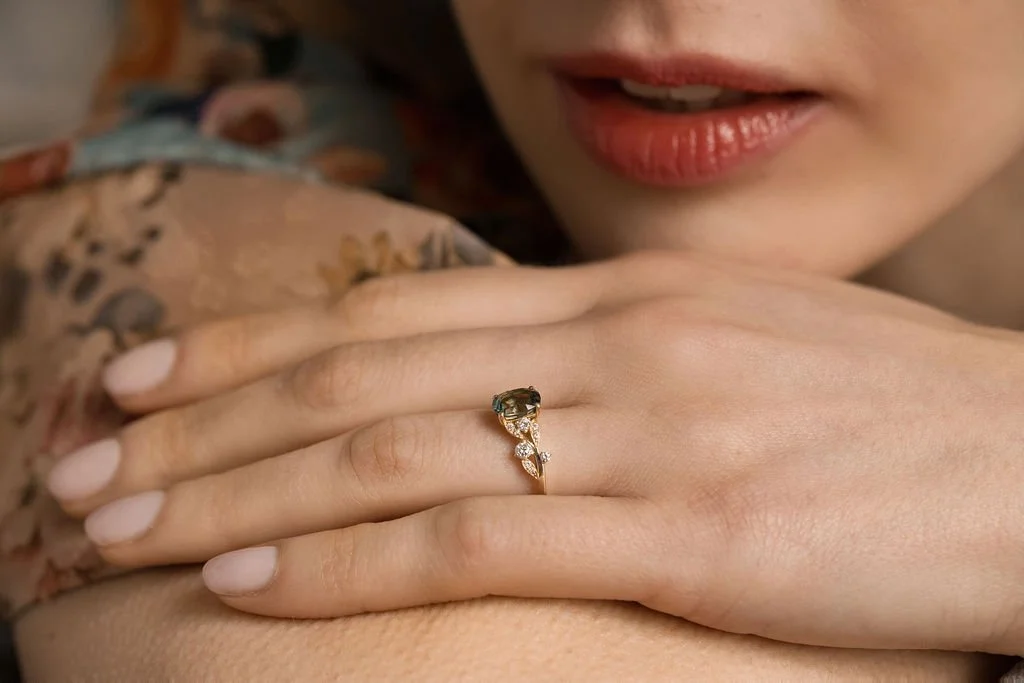 Une femme montre une bague avec un grand pierre précieuse sur son doigt. La photo est prise de près, mettant en valeur la main et le visage partiel de la femme avec des lèvres maquillées.