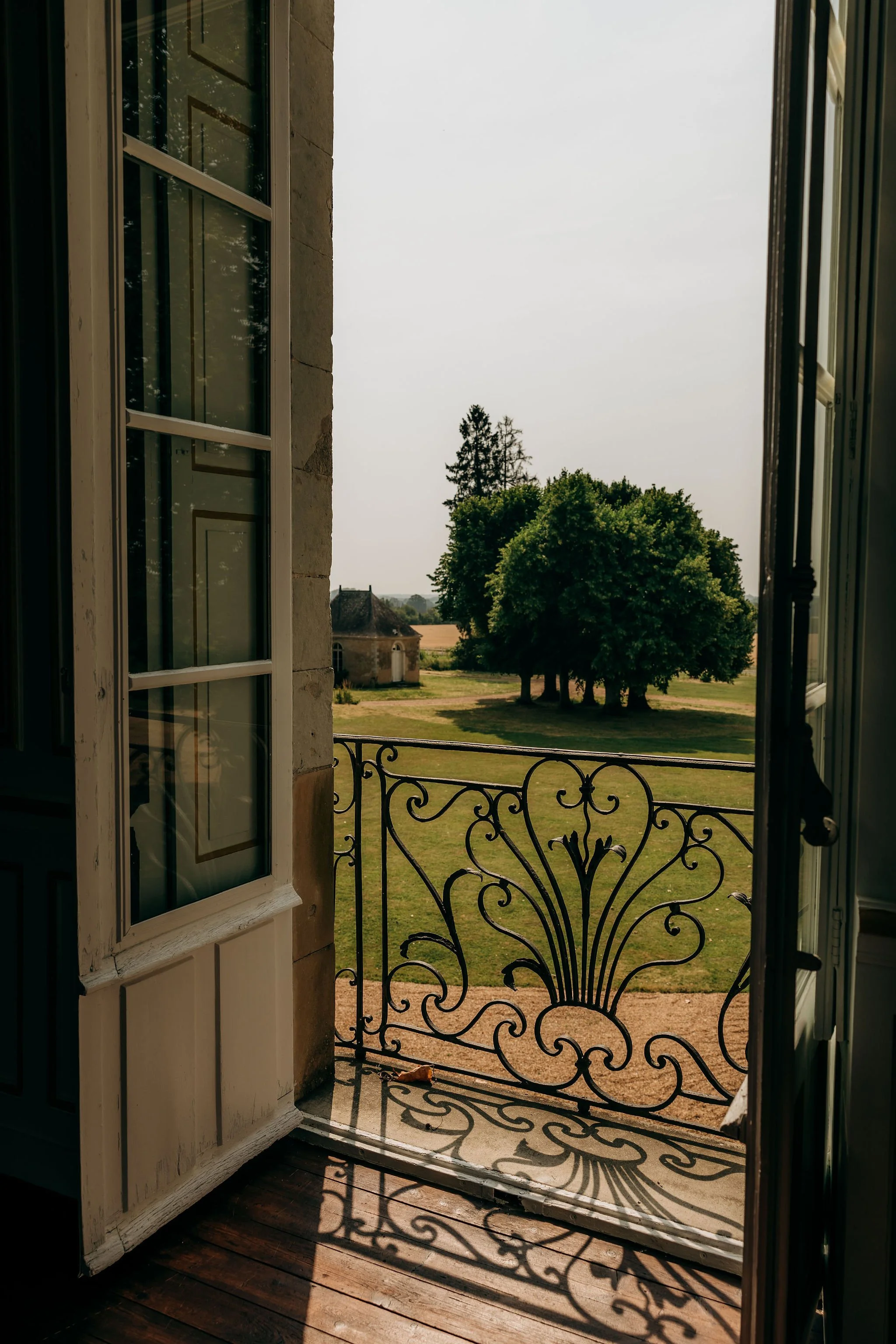 Vue depuis une fenêtre ouverte avec une porte en fer forgé, montrant un paysage de champs et d'arbres sous un ciel dégagé.