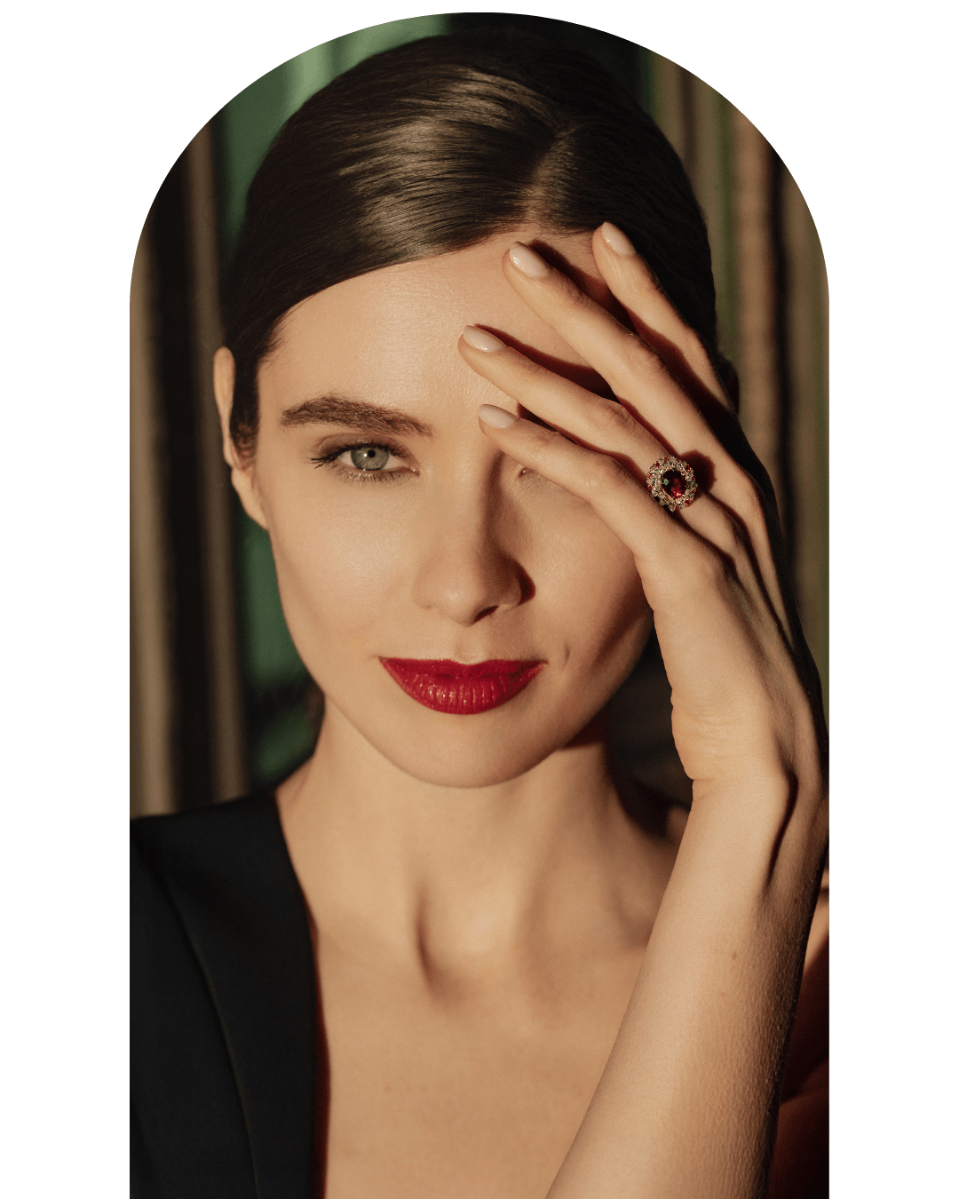 Une femme avec les cheveux bruns, maquillage rouge à lèvres, portant une bague orné de pierres rouges, pose la main sur son visage, presque couvrant un œil.