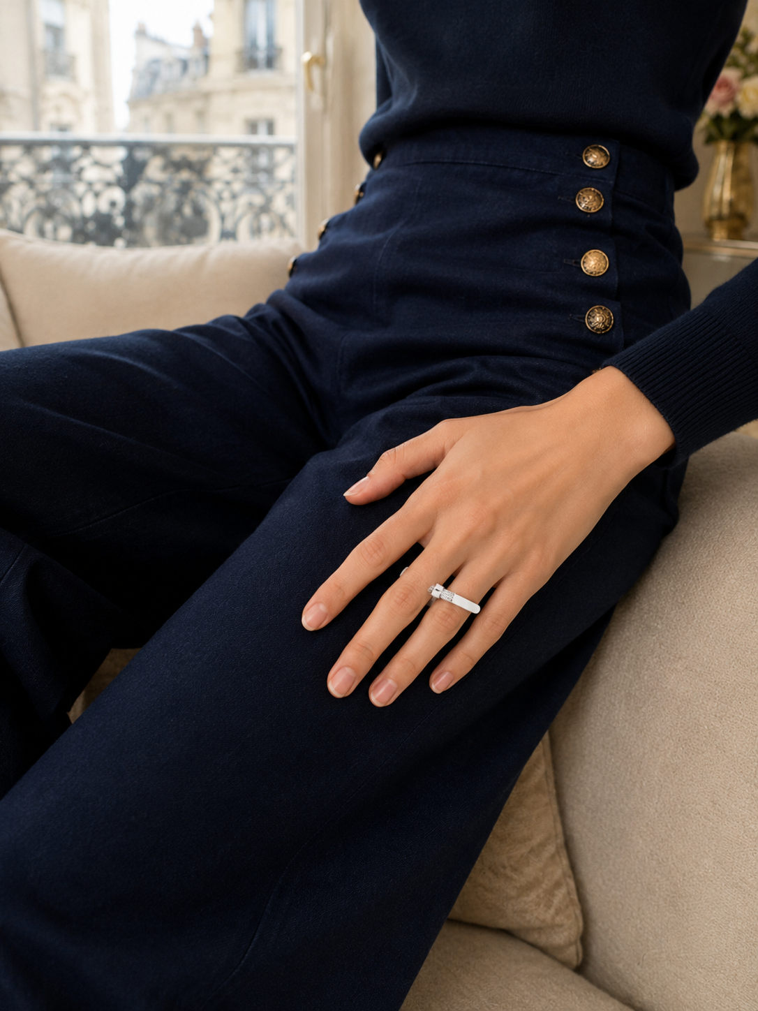 Une femme portant un haut noir et un pantalon en velours bleu foncé, avec des boutons en or sur la taille, assise sur un canapé avec une fenêtre donnant sur un bâtiment historique parisien en arrière-plan. Elle porte une bague en diamant sur son doigt.