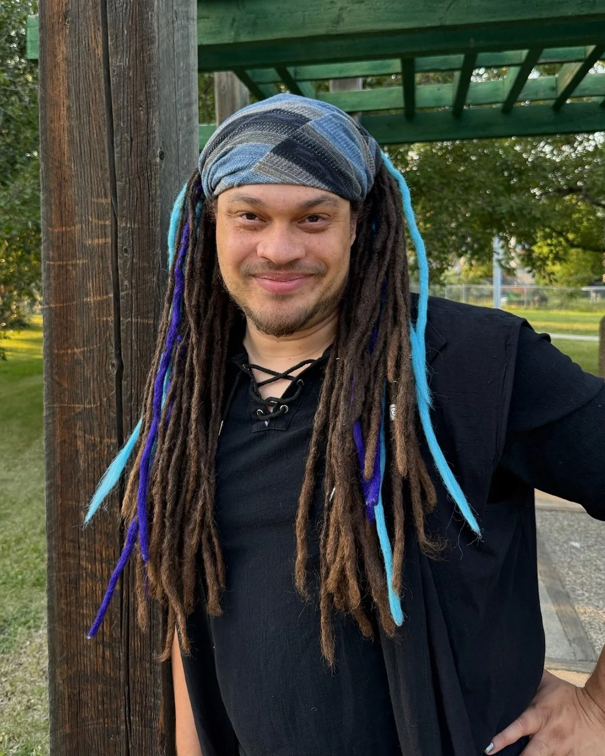 Meric Moir, a man with brown and blue dreadlocks leaning against a wooden post and smiling at the camera.