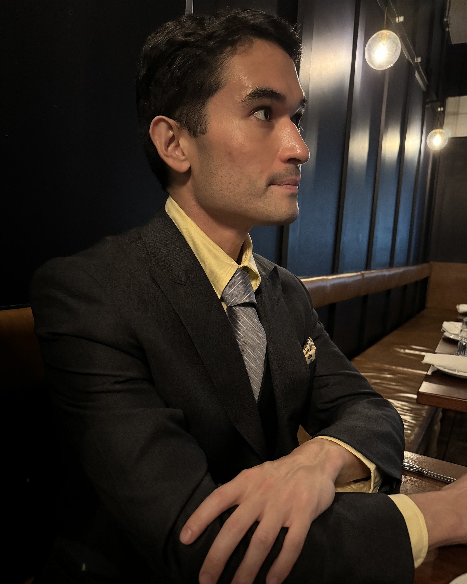 James Mendex Hodes, a dark olive-skinned man wearing a black suit with a yellow button down shirt. He's looking away from the camera.