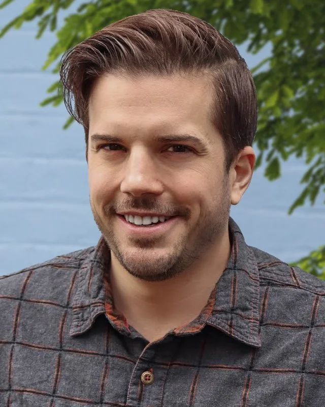 Andrew Fischer, a light skinned man with short brunette hair, wearing a grey, flannel, button down shirt and smiling at the camera.