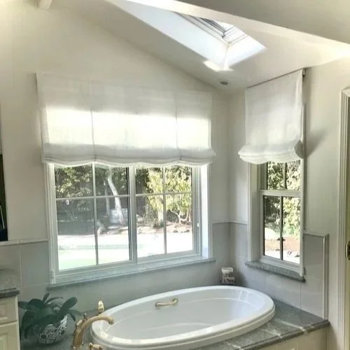 Bright bathroom with a large bathtub under three windows, white Roman shades, and a skylight overhead revealing trees outside.