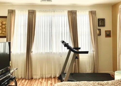 Living room with large window, sheer curtains, and beige drapes, featuring a treadmill facing the window, a TV on a stand, and framed pictures on the wall.