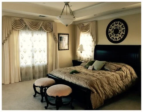 Bedroom with window curtains, a bed with pillows and a blanket, a nightstand with a lamp, wall clock, and two upholstered stools in front of the bed.
