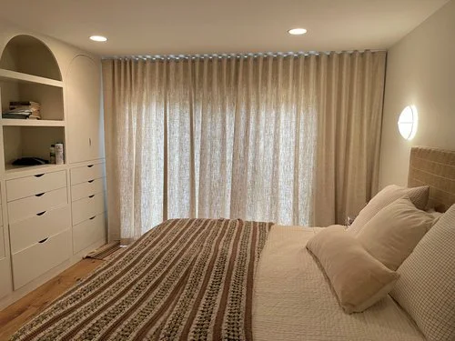 A neatly made bed with white pillows in a bedroom with beige curtains covering a window, a wall-mounted light, and white built-in shelves.
