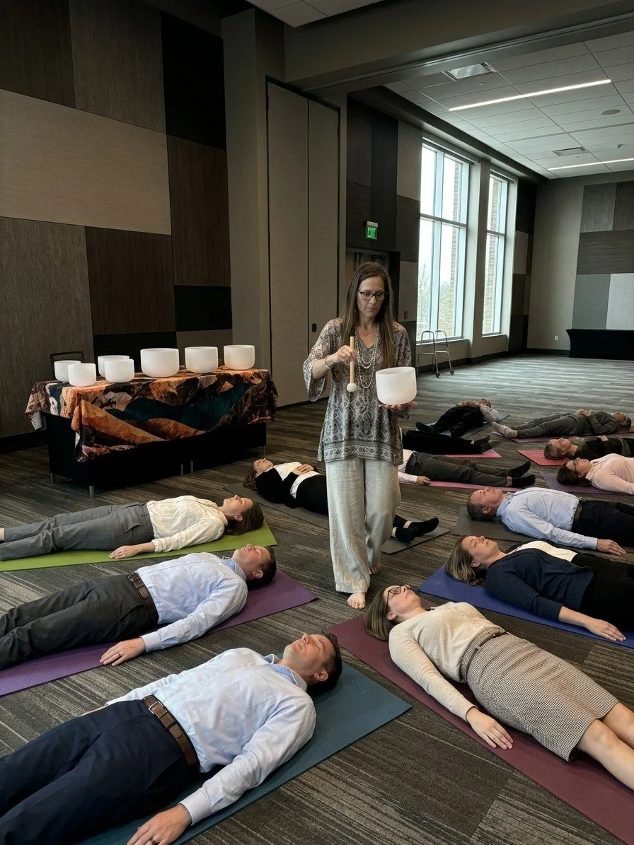 A woman leading a sound healing or meditation session using singing bowls in a spacious, well-lit room with large windows. Several participants are lying on yoga mats on the floor, relaxing with eyes closed.