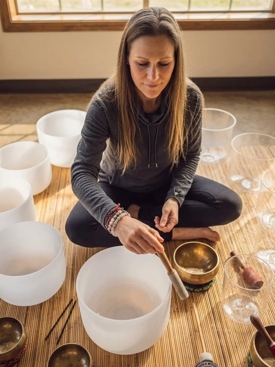 A woman sitting cross-legged on a wooden mat, playing crystal singing bowls with a mallet, surrounded by other bowls, in a well-lit indoor space.