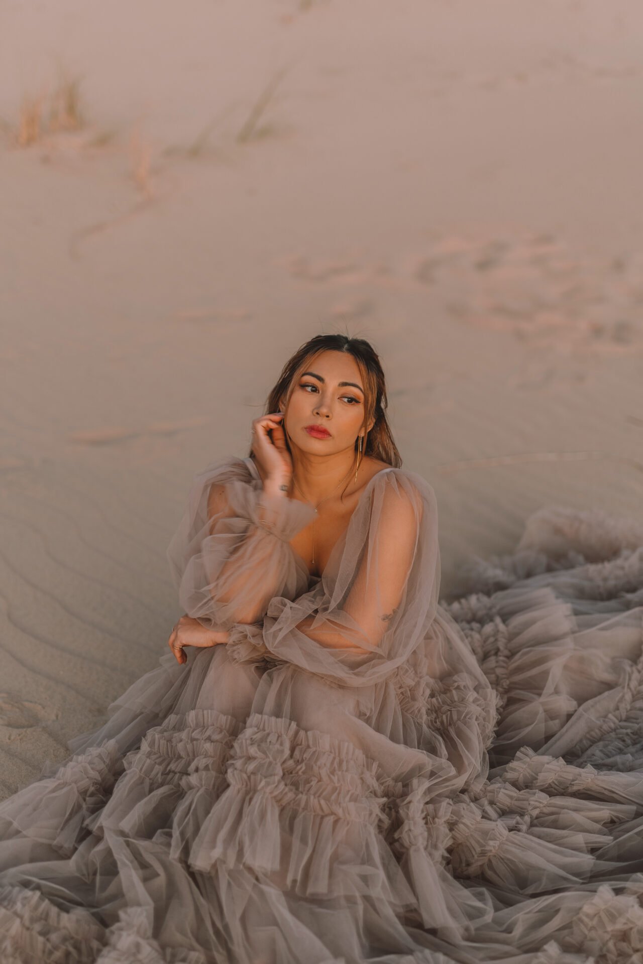 A woman wearing a flowing, ruffled, beige dress sitting on sandy ground with a contemplative expression